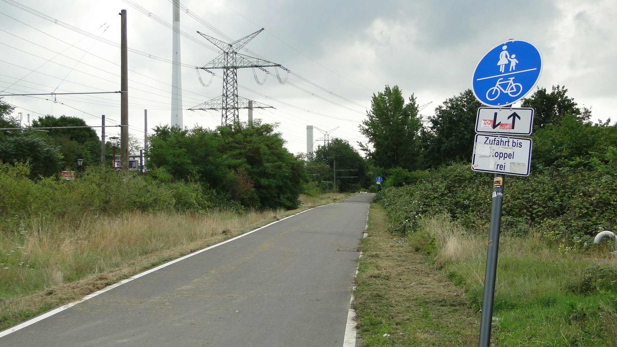 An einem asphaltierten Radweg entlang einer Schienentrasse steht ein Schild, das den Weg als Rad- und Fußweg ausweist.