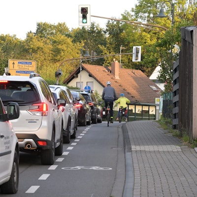 Autos vor einer Ampel, rechts ein Rad-Schutzstreifen mit zwei Fahrradfahrern