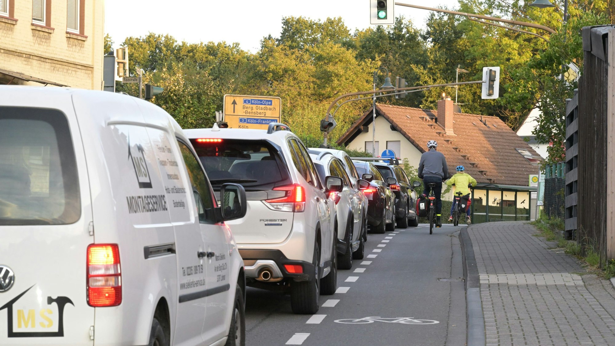 Autos vor einer Ampel, rechts ein Rad-Schutzstreifen mit zwei Fahrradfahrern