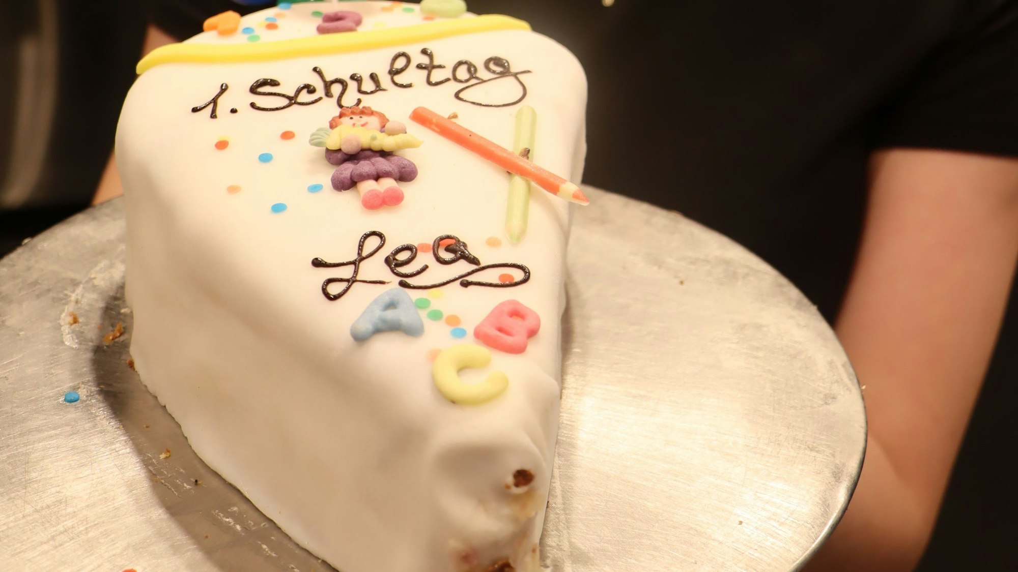 Zu sehen ist eine Nahaufnahme eines Tortenstücks mit weißer Glasur, auf dem steht: 1. Schultag Lea. Dekoriert ist die Torte mit bunten Zuckerbuchstaben und Stiften.