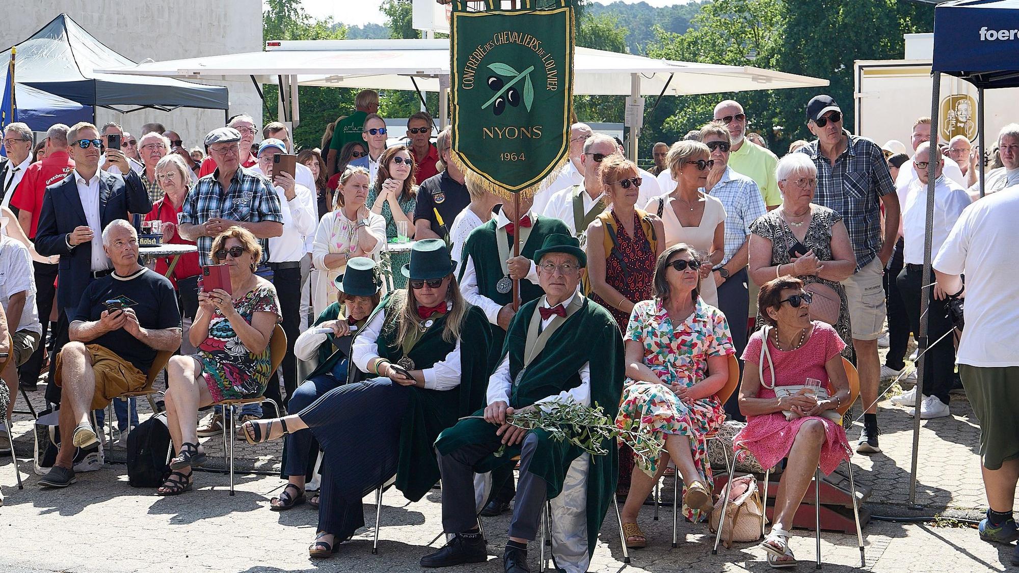 Drei neue Ritter sitzen mit Zylindern und dunklen Umhängen auf Stühlen unter einer Fahne. Dahinter stehen Festbesucher.