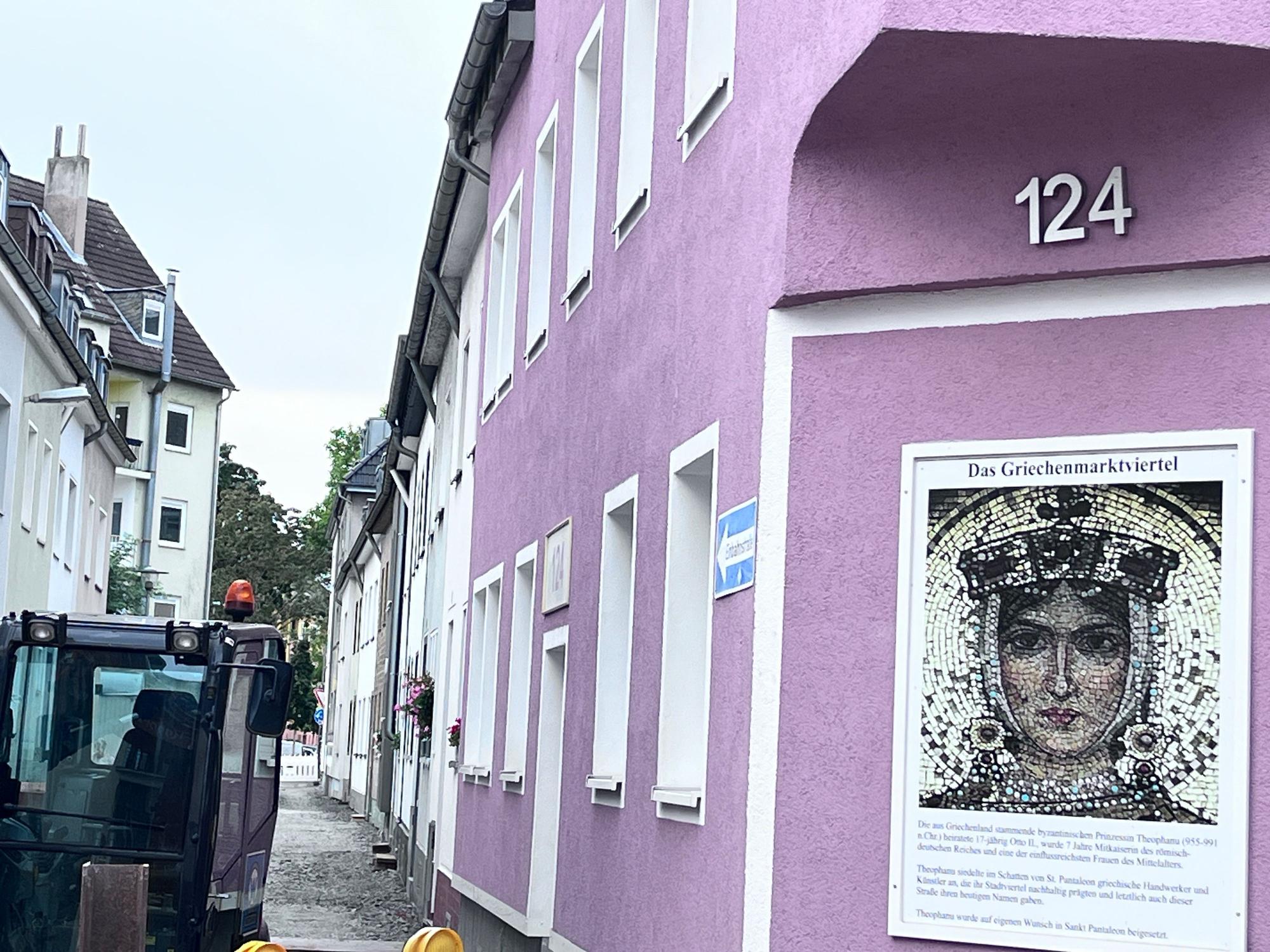 Eine kleine Gasse, in denen gerade Bauarbeiten stattfinden, ist zu sehen. Im Vordergrund hängt eine Erklärtafel an einem Haus mit lilafarbener Fassade zum Namen Griechenmarktviertel.