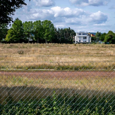 Das Bild zeigt ein hoch mit Gras bewachsenes Gelände hinter einem Maschendrahtzaun, im Hintergrund Wohnhäuser und Bäume.