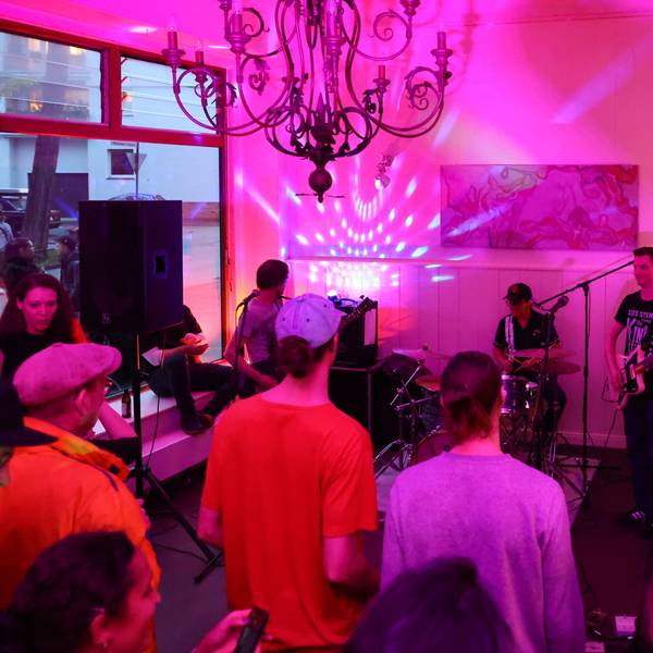 In einem Café spielt eine Band, Menschen stehen im leergeräumten Gastraum und hören zu.