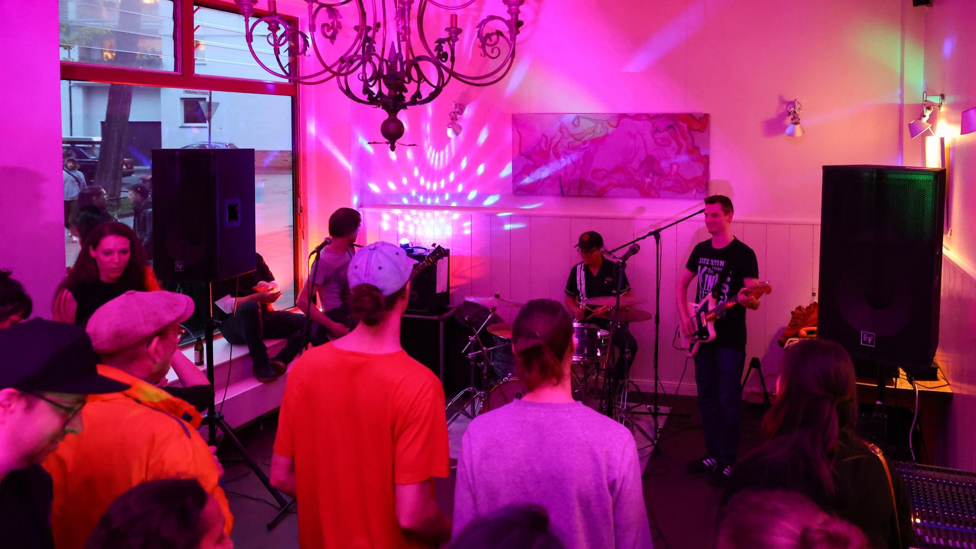 In einem Café spielt eine Band, Menschen stehen im leergeräumten Gastraum und hören zu.