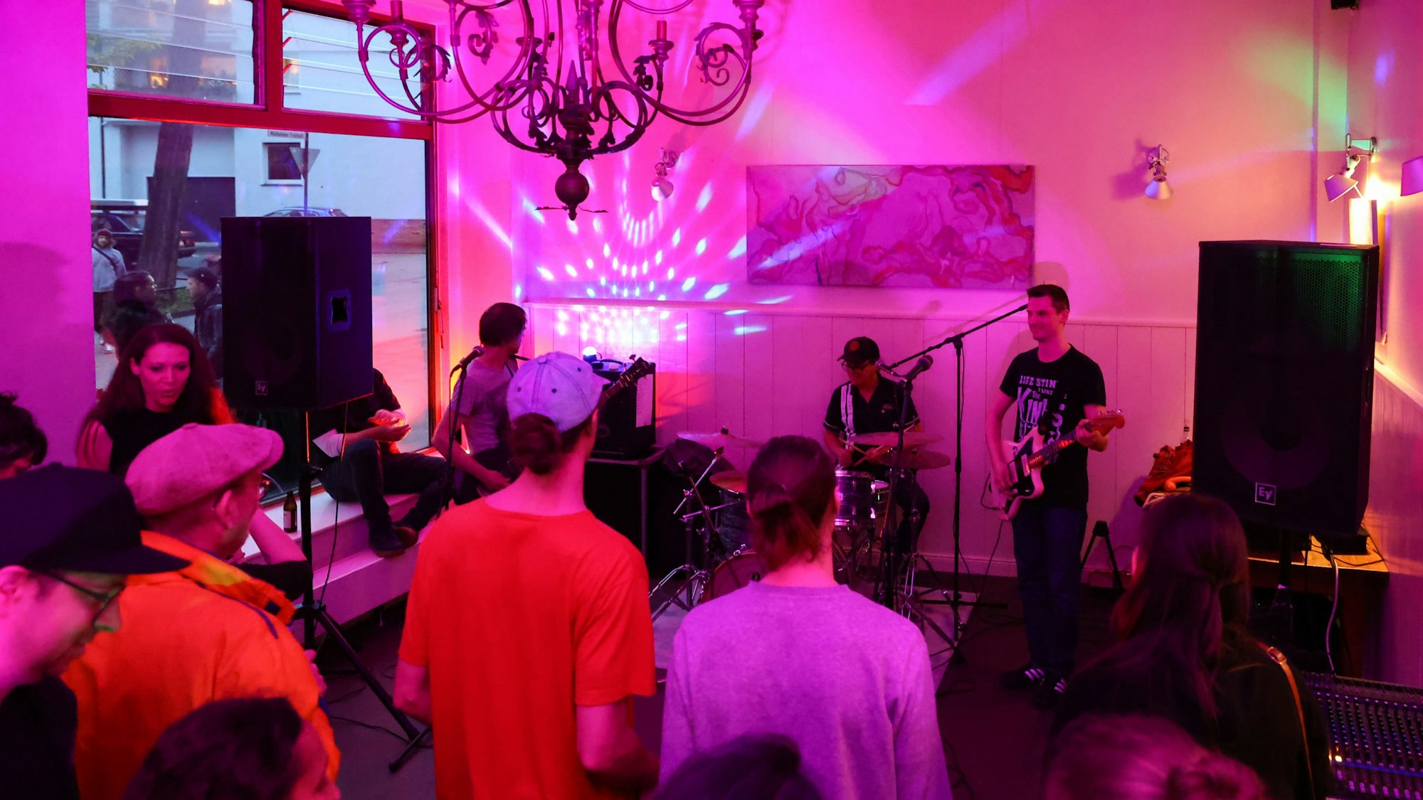 In einem Café spielt eine Band, Menschen stehen im leergeräumten Gastraum und hören zu.