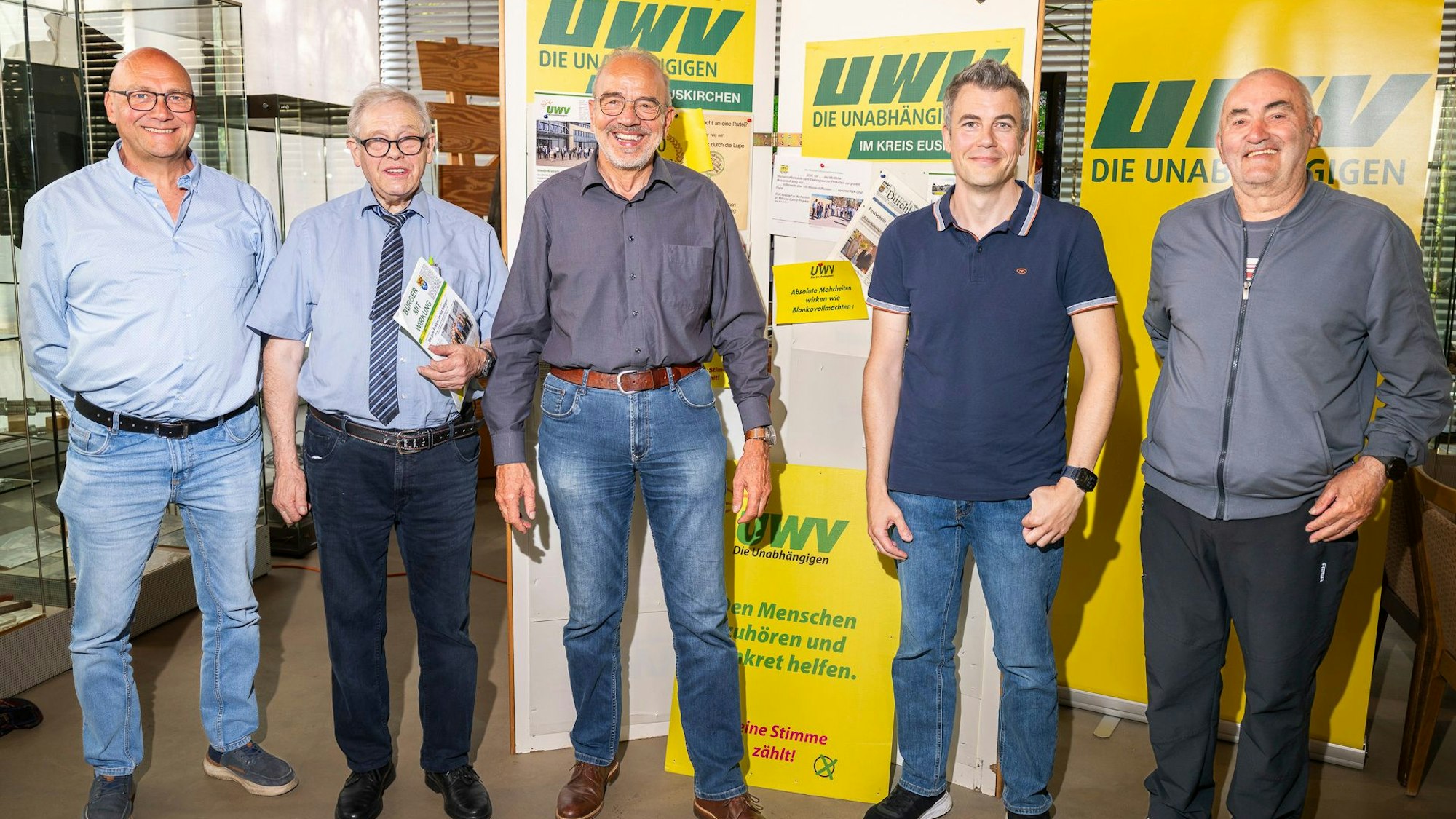 Fünf Männer haben sich vor den gelben Plakaten der UWV in einer Reihe zum Gruppenbild aufgestellt.