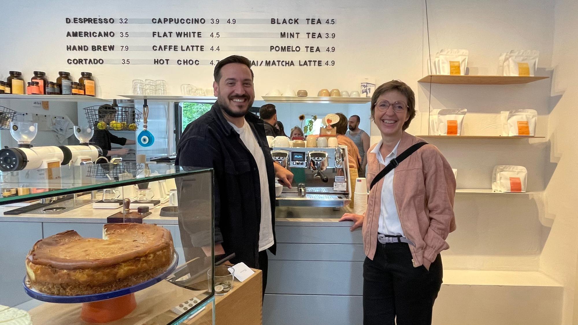 Der Betreiber des Cafés steht neben einer Frau mit Kurzhaarfrisur, die einen lachsfarbenen Blouson trägt.