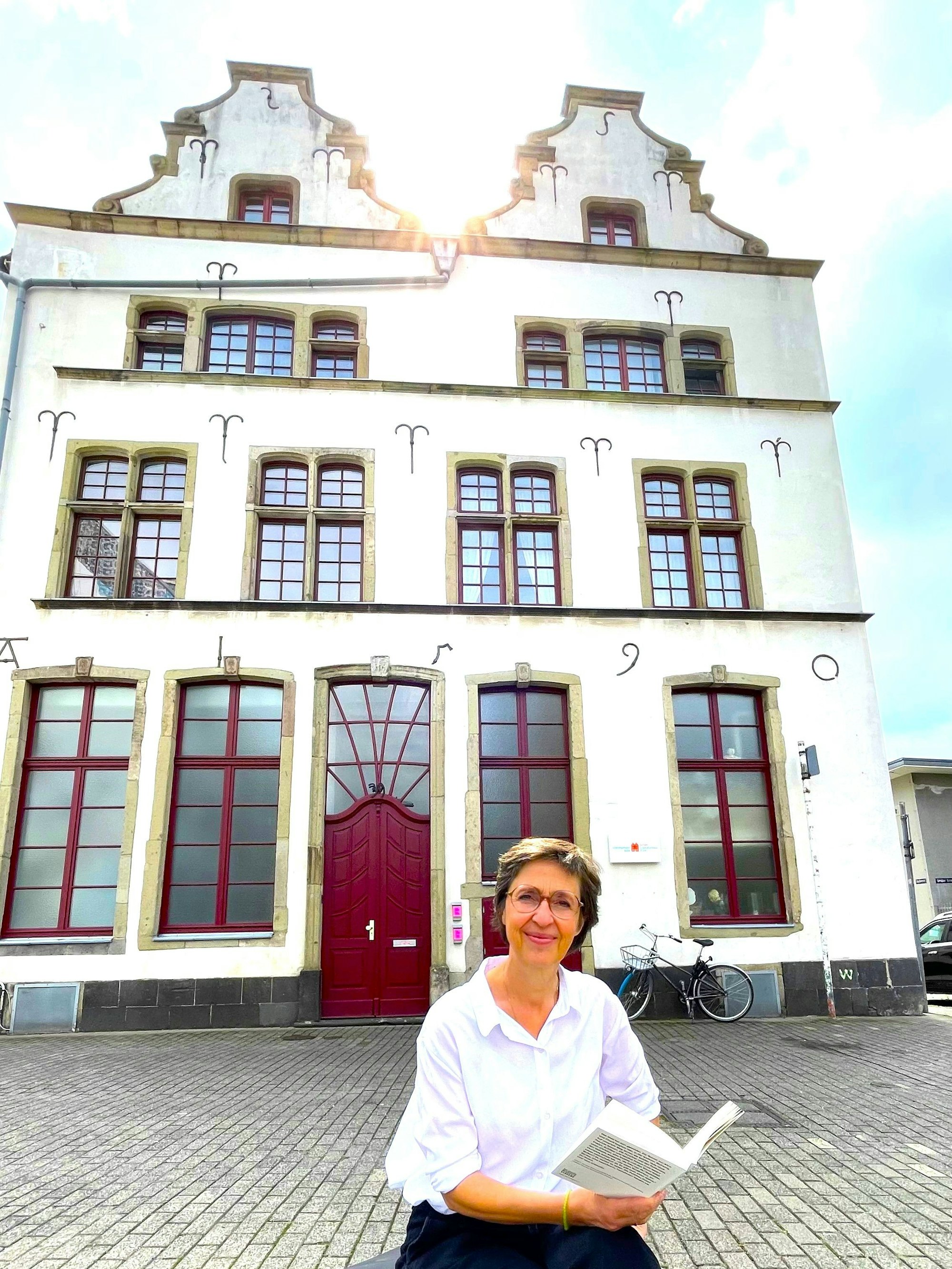 Eine Frau sitzt lesend vor einem Haus mit historischer Fassade.