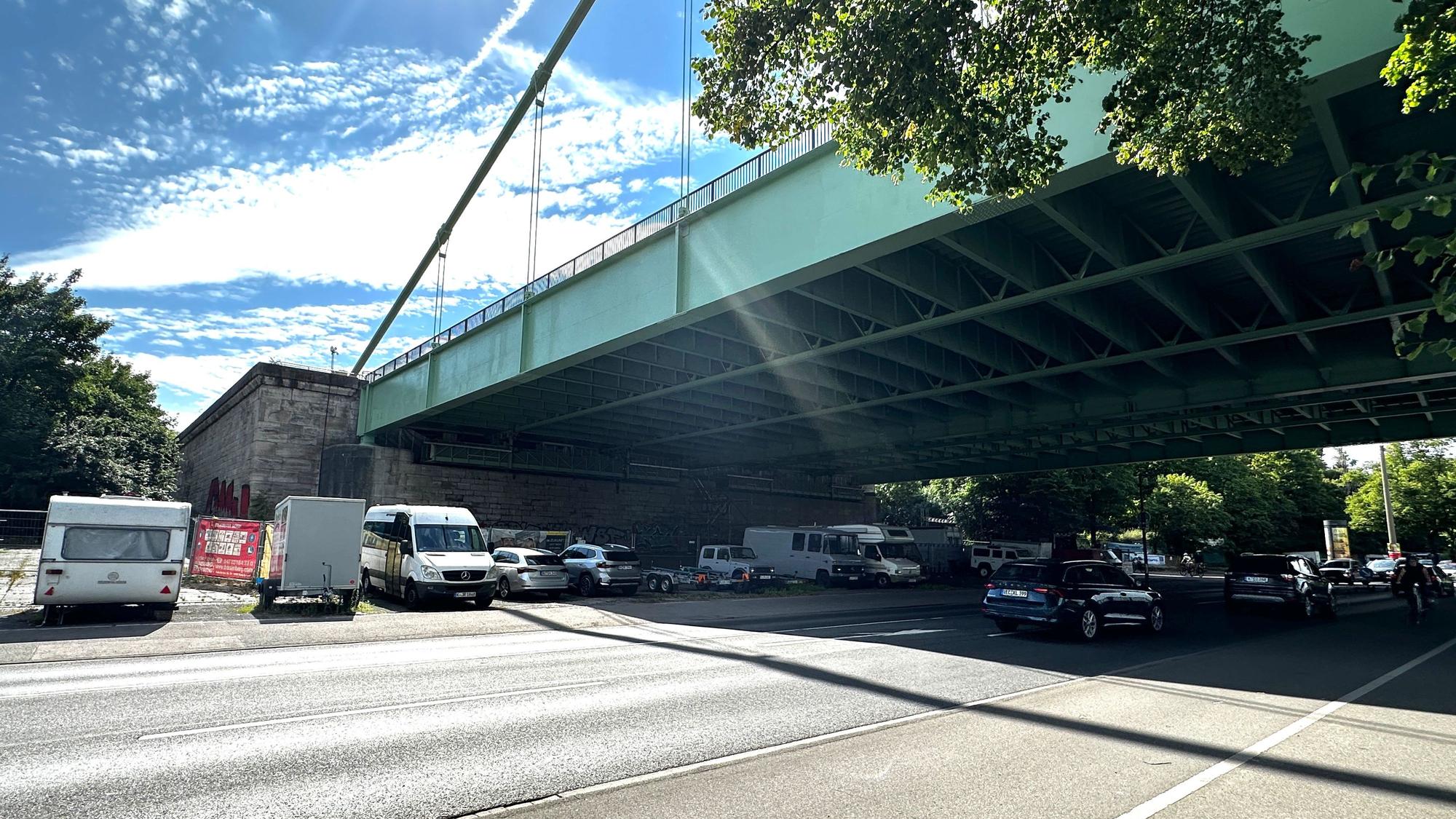 Unter einer großen Straßenbrücke verläuft eine mehrspurige Straße, neben der Straße parken einige Autos und Campingwagen unter der Brücke.
