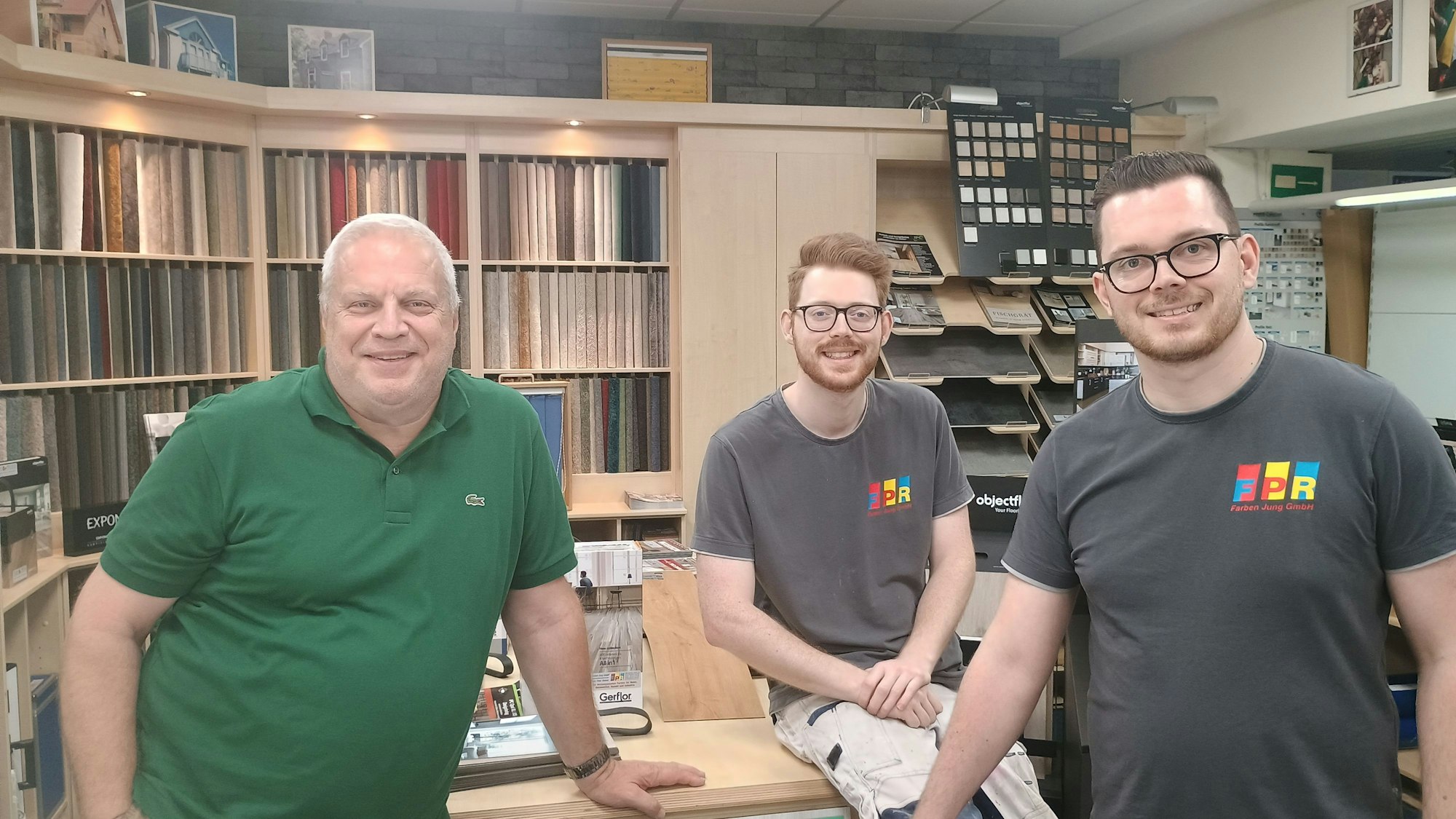 Der Inhaber steht neben seinen beiden Söhnen, die schwarze T-Shirts mit dem bunten Logo von Farben Jung. Die beiden stehen im Geschäft, im Hintergrund sind zahlreiche Exemplare für Bodenbeläge zu sehen.