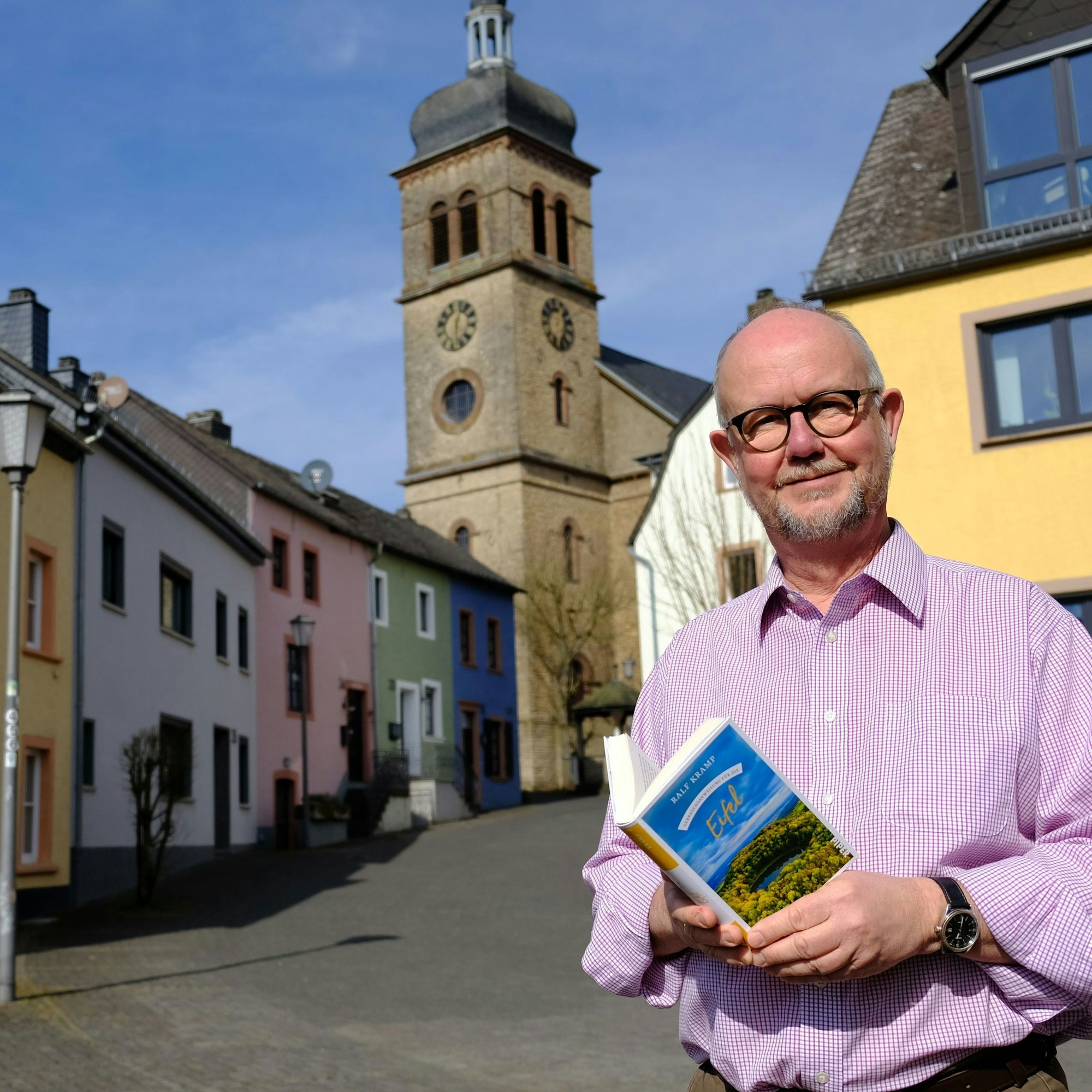 Der Schriftsteller Ralf Kramp hält sein Buch "Gebrauchsanweisung für die Eifel" in der Hand. Er steht vor einem Altstadt-Ensemble mit bunt angestrichenen Häusern in Hillesheim, im Hintergrund ragt der Turm der Pfarrkirche St. Martin empor.