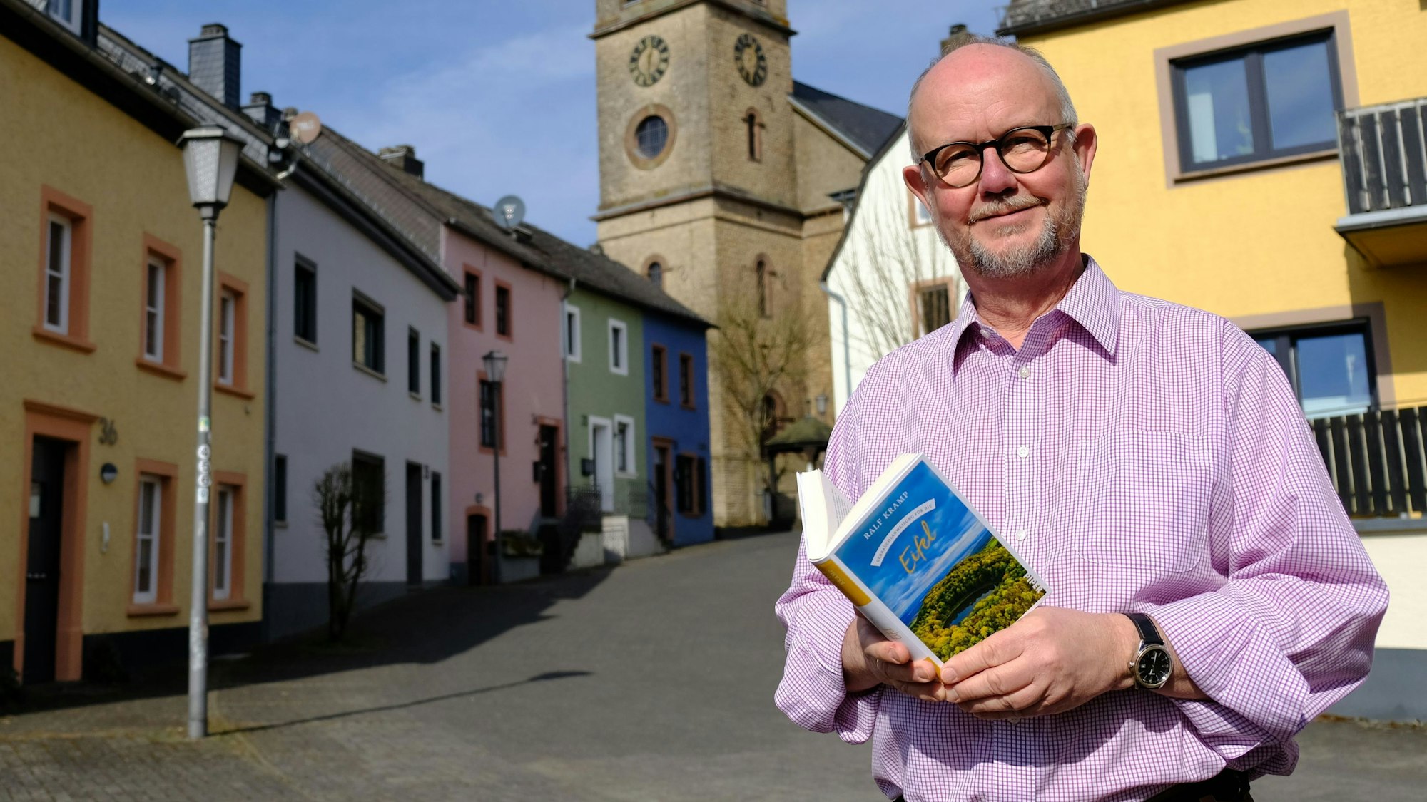 Der Schriftsteller Ralf Kramp hält sein Buch ‚Gebrauchsanweisung für die Eifel‘ in der Hand. Er steht vor einem Altstadt-Ensemble mit bunt angestrichenen Häusern in Hillesheim, imm Hintergrund ragt der Turm der Pfarrkirche St. Martin empor.
