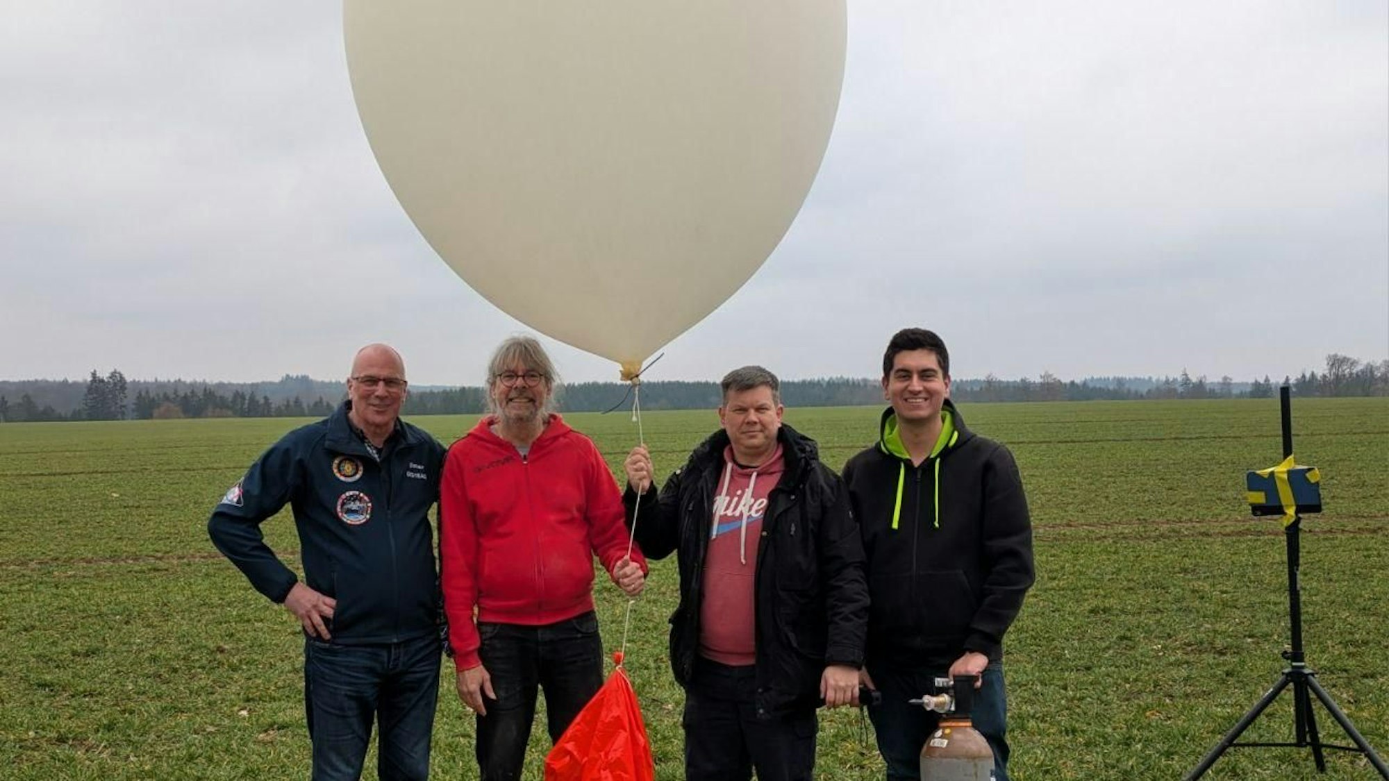 Dieter Eggert Hinsch (v.l.), Martin Kopp, Denis Apel und Manuel Bülo stehen auf einer Wiese, sie haben einen Ballon und eine Gasflasche dabei.