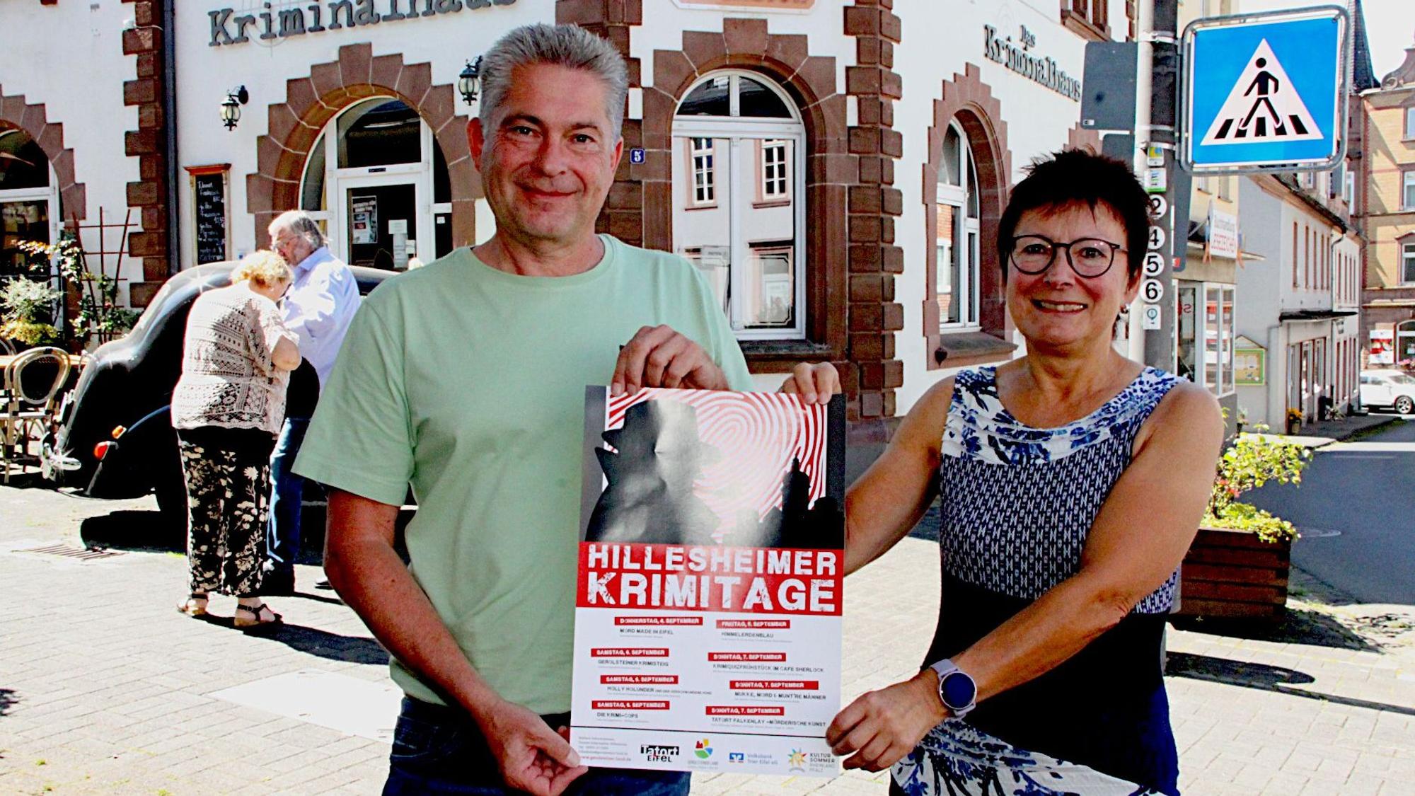 Frank Reuter und Rita Kallenberg stehen vor dem „Kriminalhaus“ und halten ein Plakat der Krimitage in den Händen.