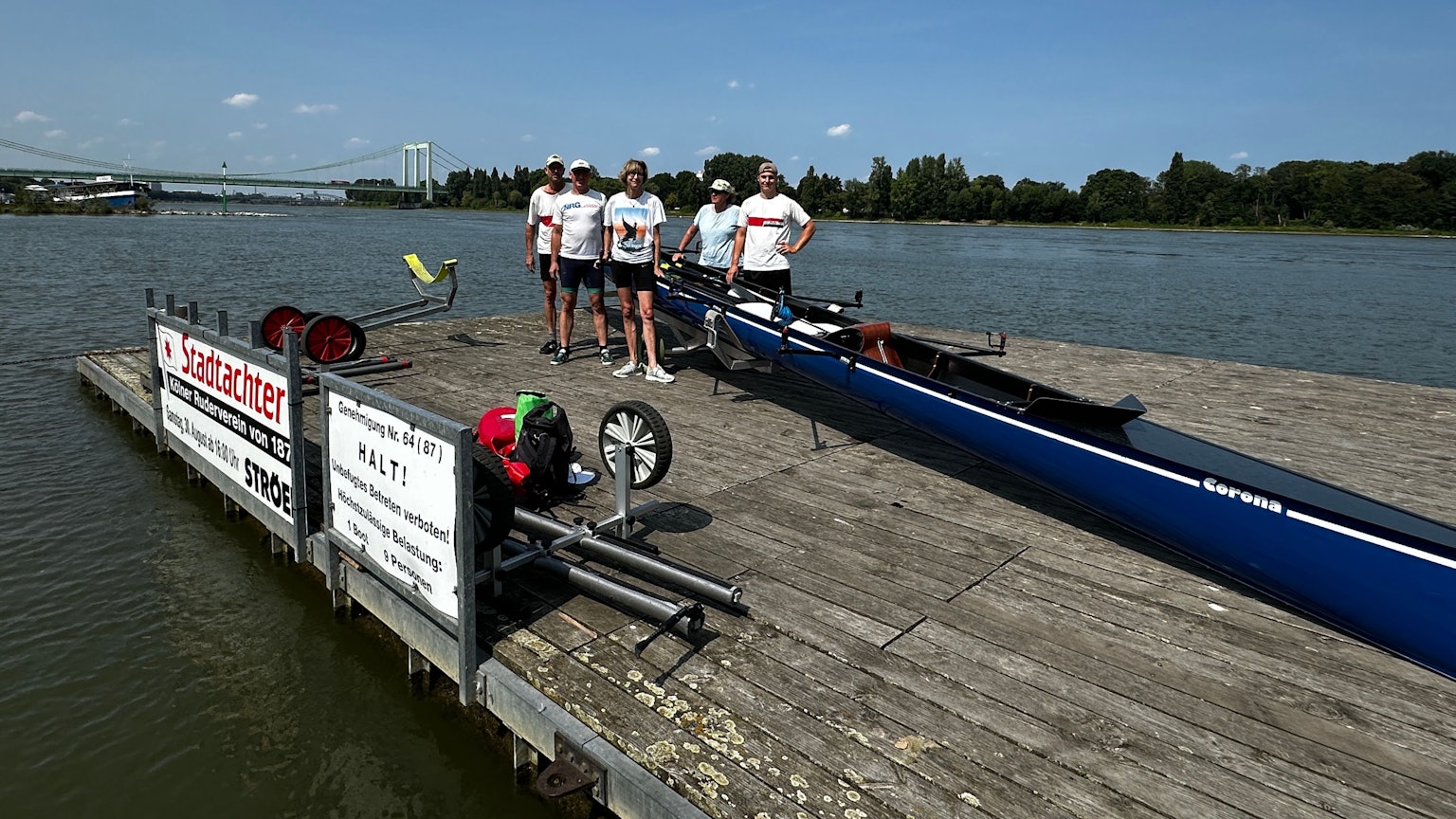 Auf einem Bootssteg stehen fünf Menschen neben einem Ruderboot, im Hintergrund sind der Rhein und die Rodenkirchener Brücke zu sehen.