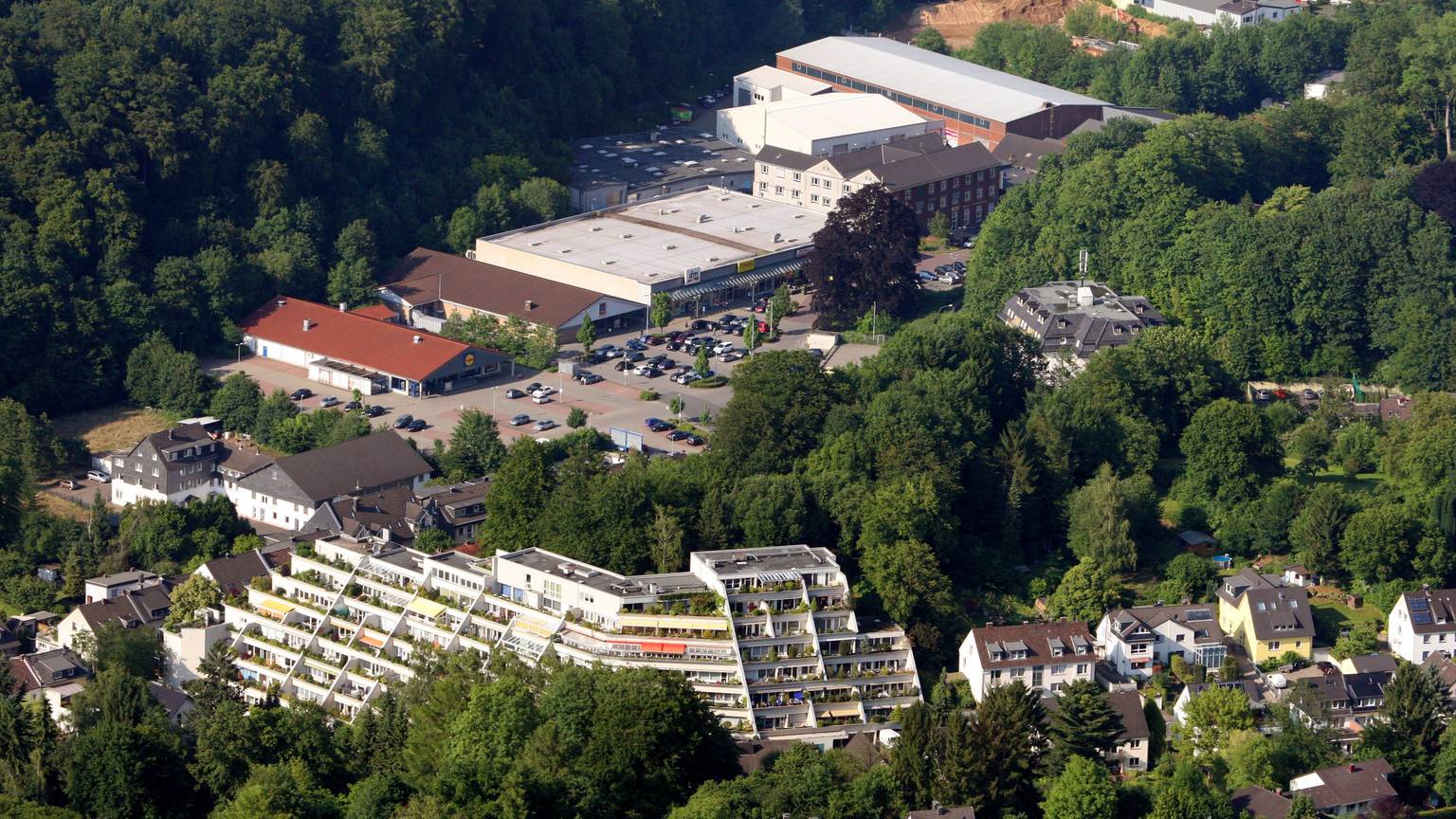 Eine Lufaufnahme mit einem terrassenartig angelegten Hochhaus, Einfamilienhäusern und einem großen Parkplatz vor einem Einkaufszentrum.