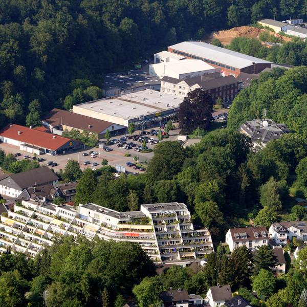 Eine Lufaufnahme mit einem terrassenartig angelegten Hochhaus, Einfamilienhäusern und einem großen Parkplatz vor einem Einkaufszentrum.