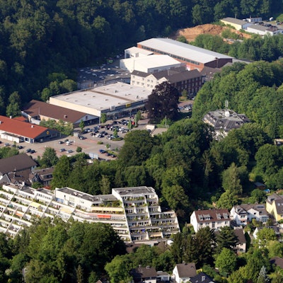Eine Lufaufnahme mit einem terrassenartig angelegten Hochhaus, Einfamilienhäusern und einem großen Parkplatz vor einem Einkaufszentrum.