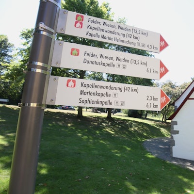 Schilder weisen den Weg für die Kapellenwanderung, im Hintergrund steht eine kleine Kapelle.