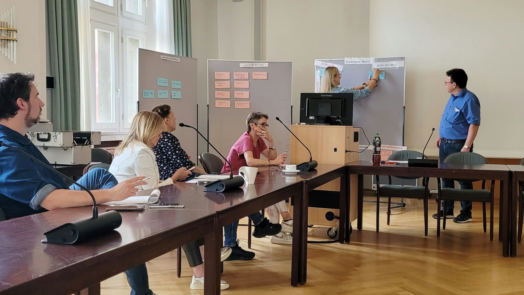 Frauen und Männer sitzen an einem Tisch. Sie hören einen Vortrag.