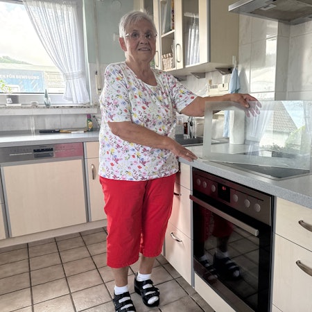 Eine ältere Dame steht in einer Küche und hält eine Glasplatte fest, die auf dem Herd steht.