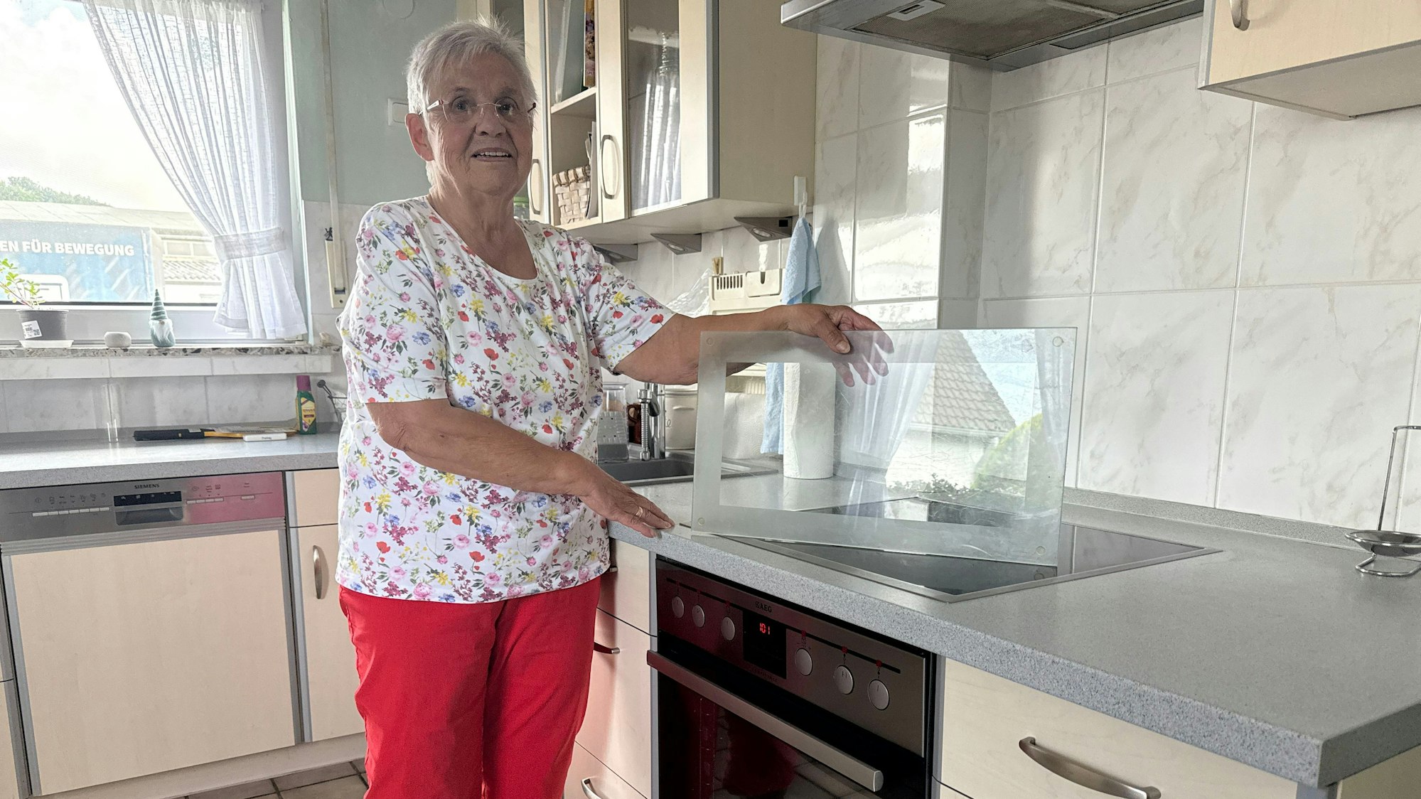 Eine ältere Dame steht in einer Küche und hält eine Glasplatte fest, die auf dem Herd steht.