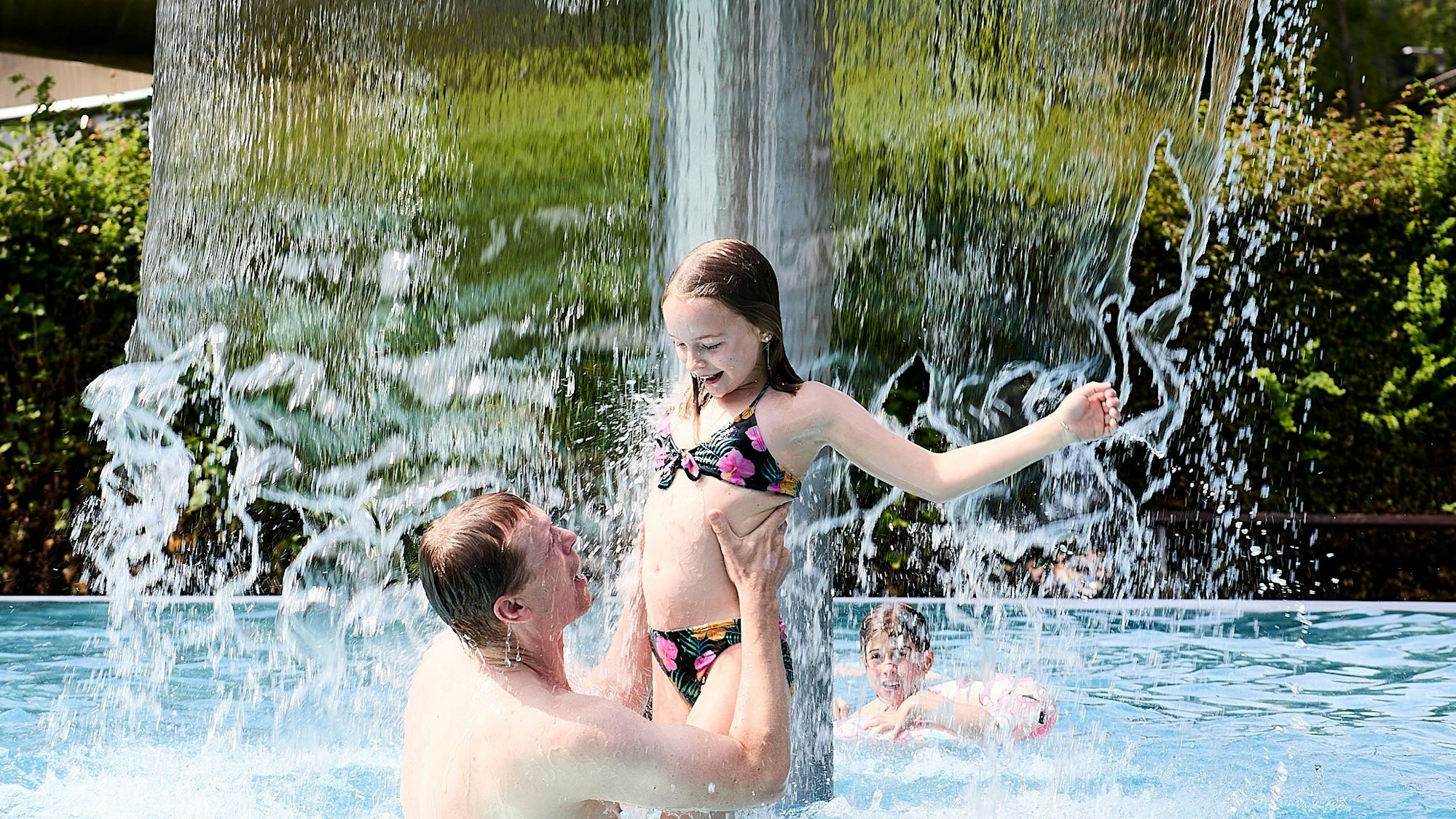 Ein Mann steht mit seiner Tochter im Freibad unter dem Wasserpilz.