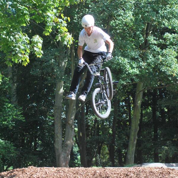 Das Bild zeigt einen BMX-Fahrer mit Schutzhelm, der aussieht, als stehe er nach einem Sprung neben seinem Fahrrad, dessen Lenker er hält, in der Luft.