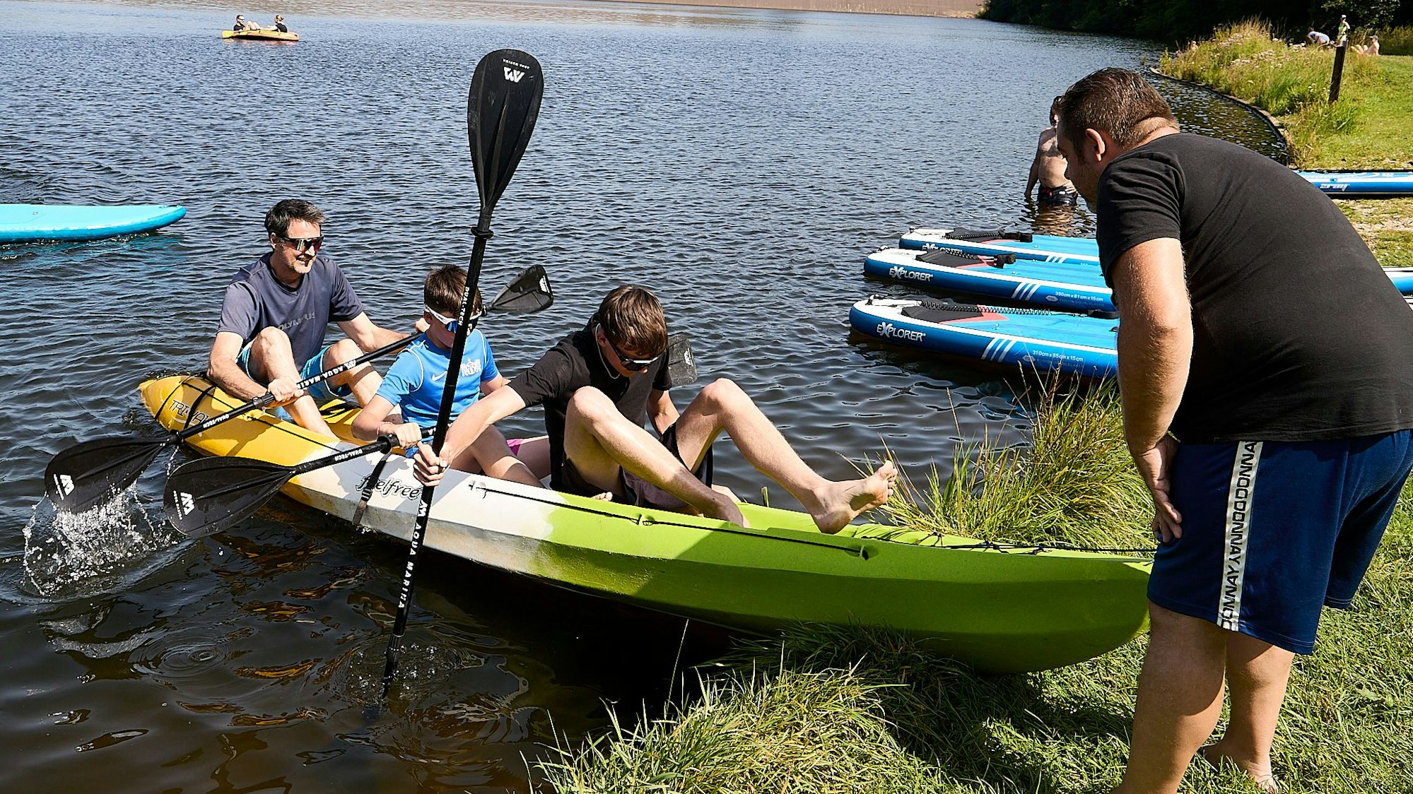 Ein Helfer im schwarzen T-Shirt schiebt ein mit drei Personen besetztes Paddelboot ins Wasser.