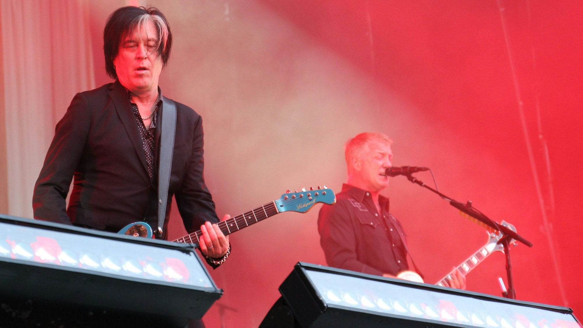 Zwei Gitarristen stehen auf einer in rotem Licht getauchten Bühne. Es sind Troy Van Leeuwen und Josh Homme von Queens Of The Stone Age.