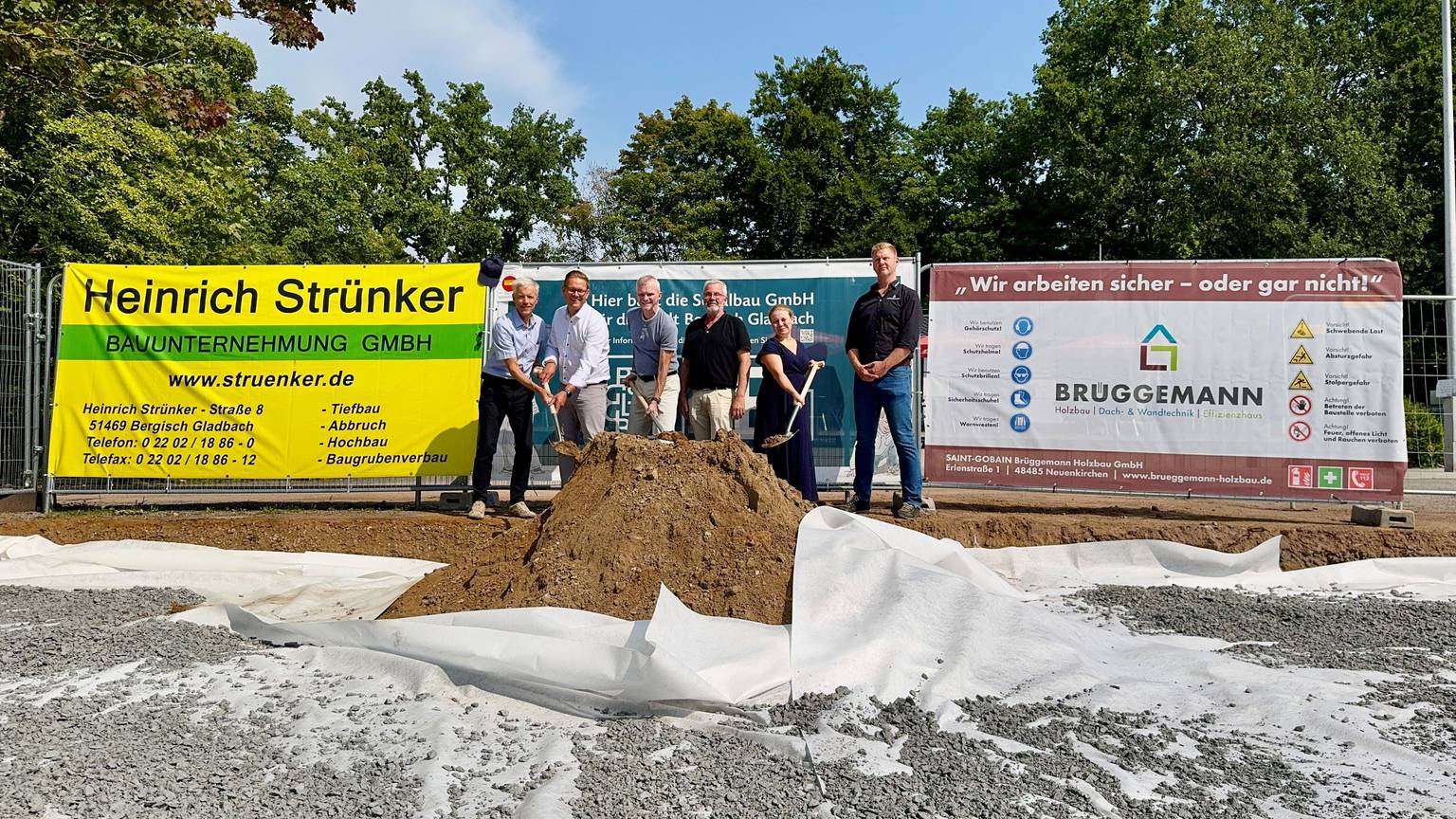 Fünf Männer und eine Frau stehen mit Spaten vor einem Haufen Erde. Dahinter Bauplakate.