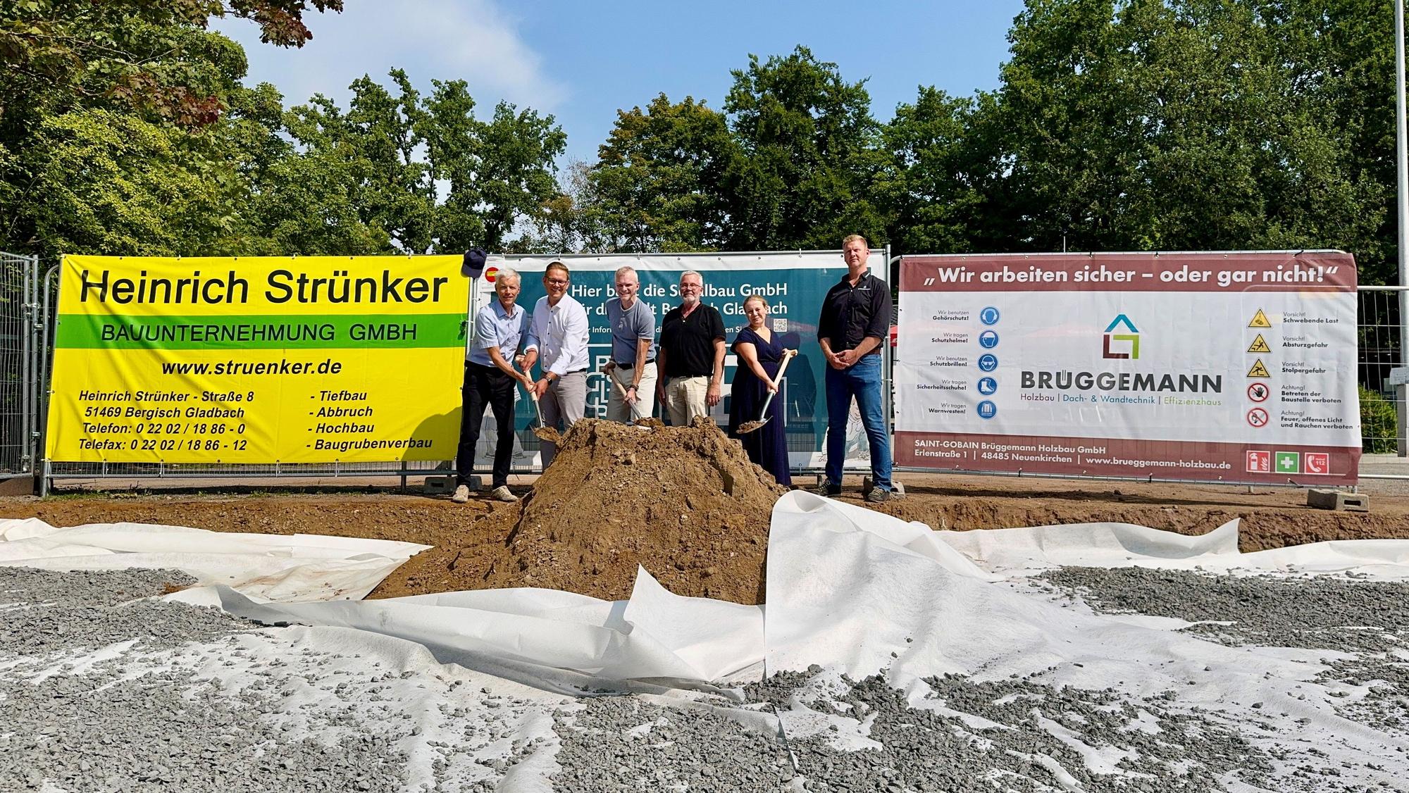 Fünf Männer und eine Frau stehen mit Spaten vor einem Haufen Erde. Dahinter Bauplakate.