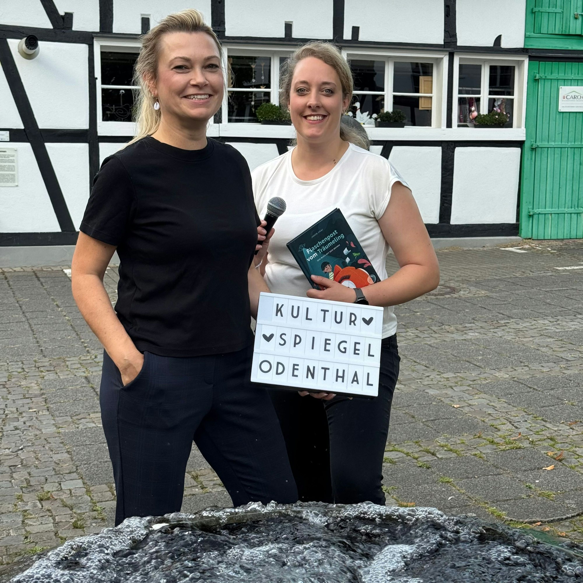 Zwei Frauen stehen vor einem Fachwerkgebäude und haben ein Schild mit der Aufschrift „Kultur Spiegel Odenthal“ in der Hand.