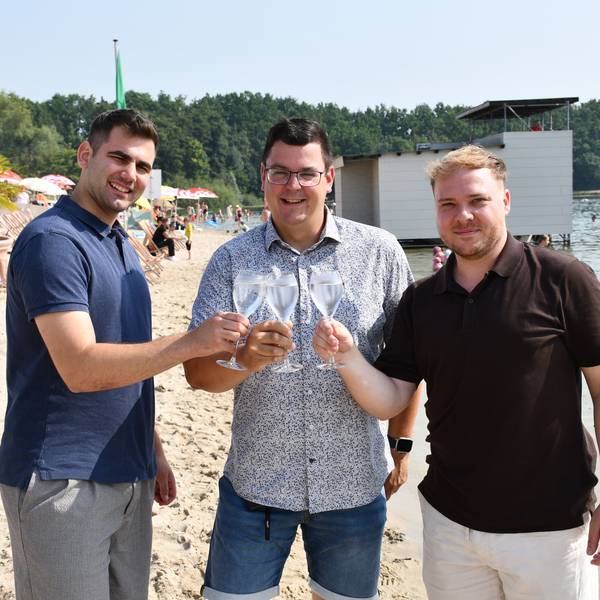 Das Foto zeigt drei Männer lockerer Kleidung im Strandbad, die mit Sektgläsern anstoßen.