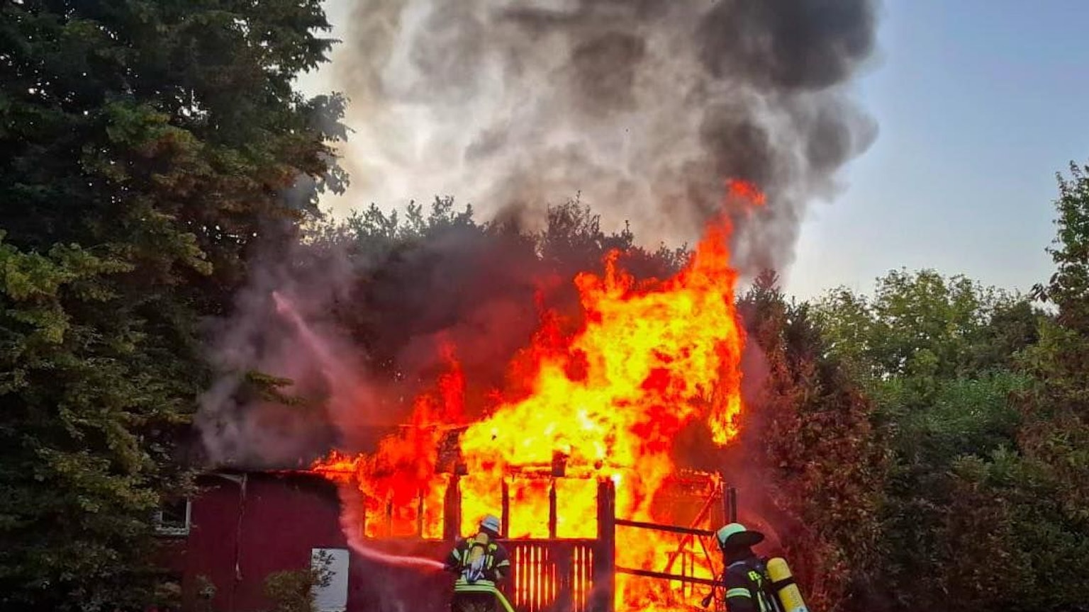 Ein Gartenhaus brennt lichterloh. Im Vordergrund Feuerwehrleute im Einsatz.