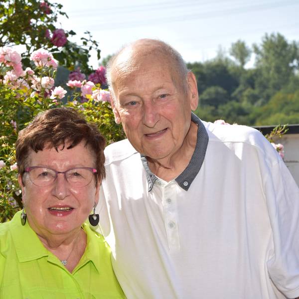 Das Paar steht vor einem Busch in voller Blühte. Anna Schiffer trägt ein grünes Hemd, Bruno Schiffer ein weißes.