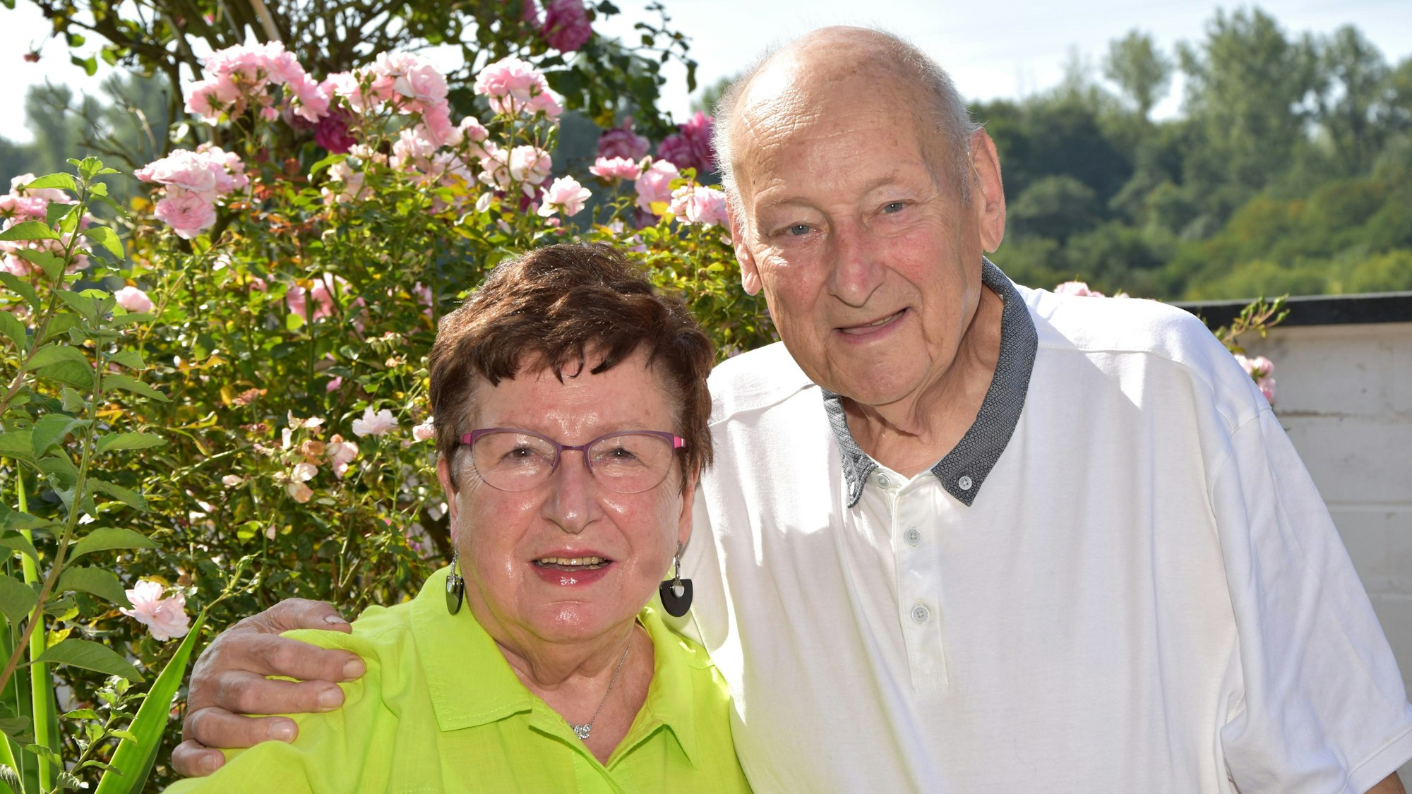 Das Paar steht vor einem Busch in voller Blühte. Anna Schiffer trägt ein grünes Hemd, Bruno Schiffer ein weißes.