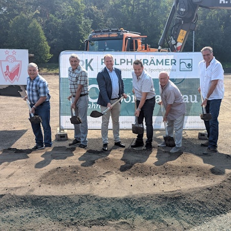 Das Foto zeigt sieben Männer mit Spaten vor einem Banner und Baumaschinen auf dem Sportplatz.