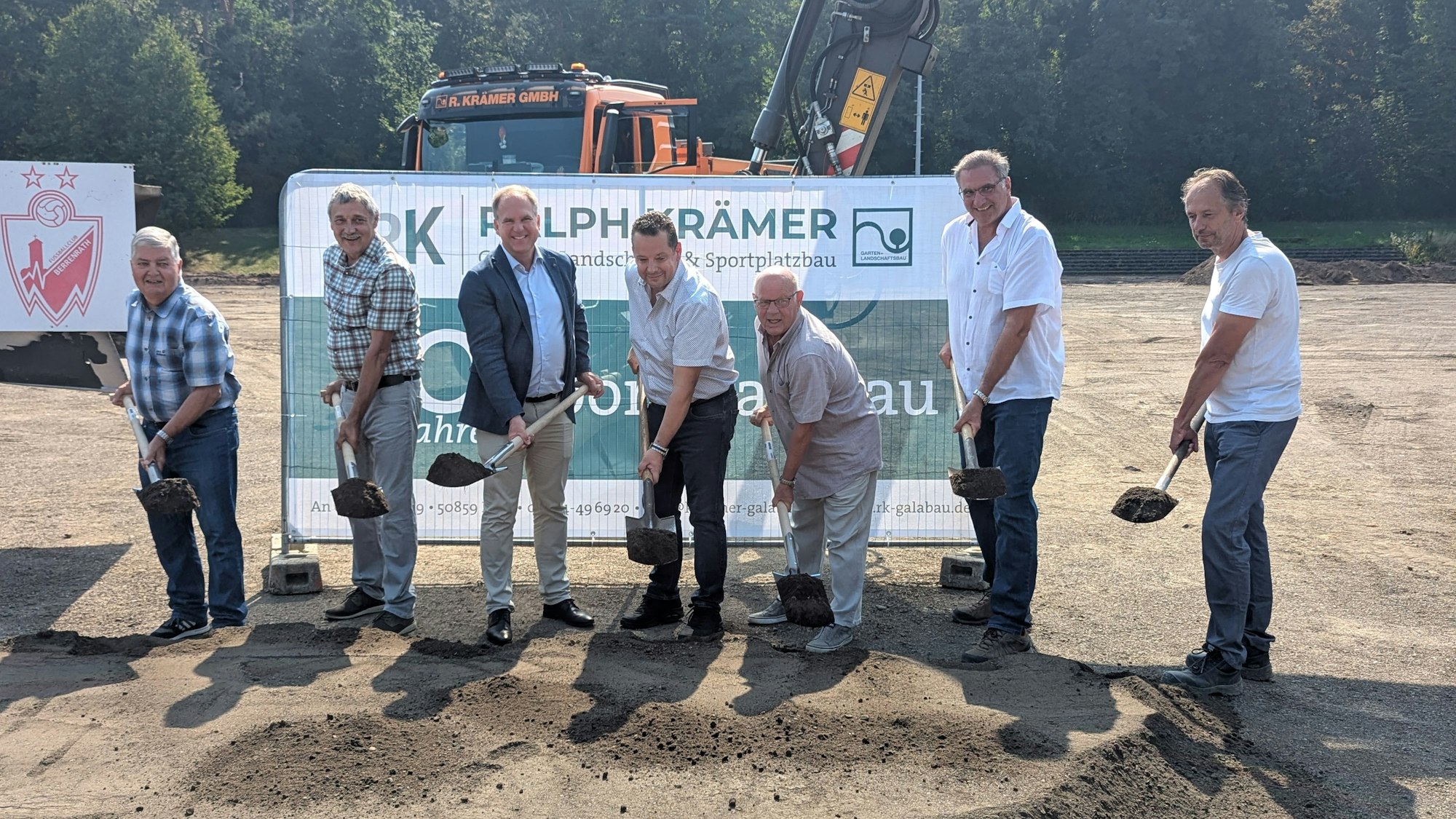 Das Foto zeigt sieben Männer mit Spaten vor einem Banner und Baumaschinen auf dem Sportplatz.