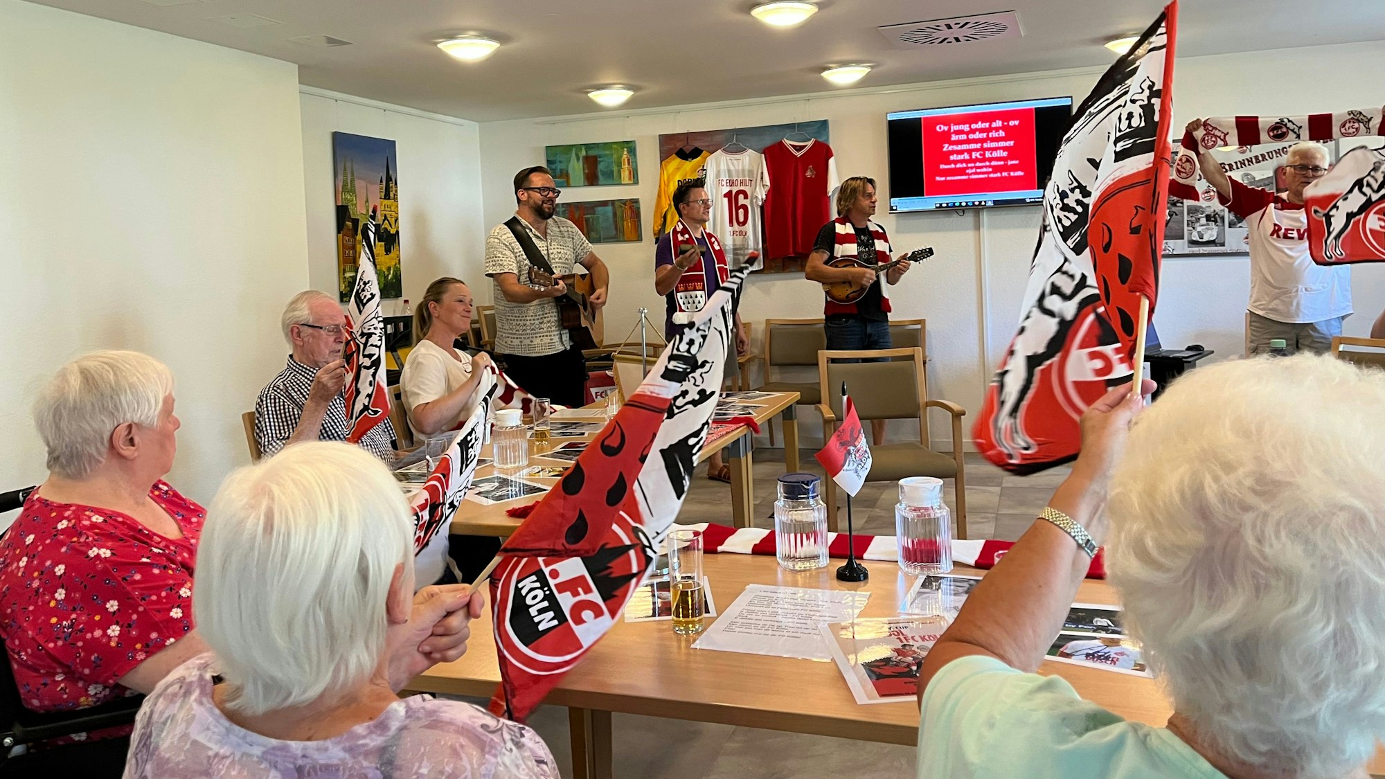Auf einem Monitor ist der Text der FC-Hymne zu sehen, Seniorinnen und Senioren schwenken FC-Fahnen und singen mit.
