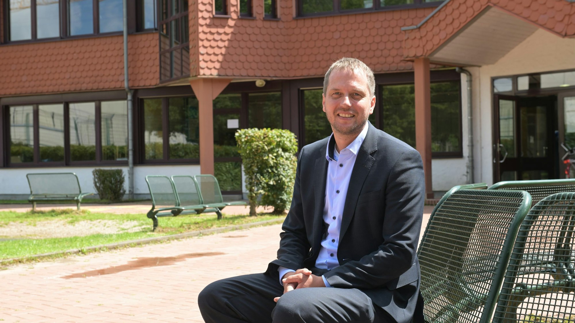 Ein Mann sitzt in einem dunklen Anzug vor einer modernen Grundschule.
