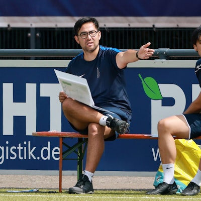 Das Foto zeigt zwei Trainer auf der Bank, der Linke streckt den Arm aus.