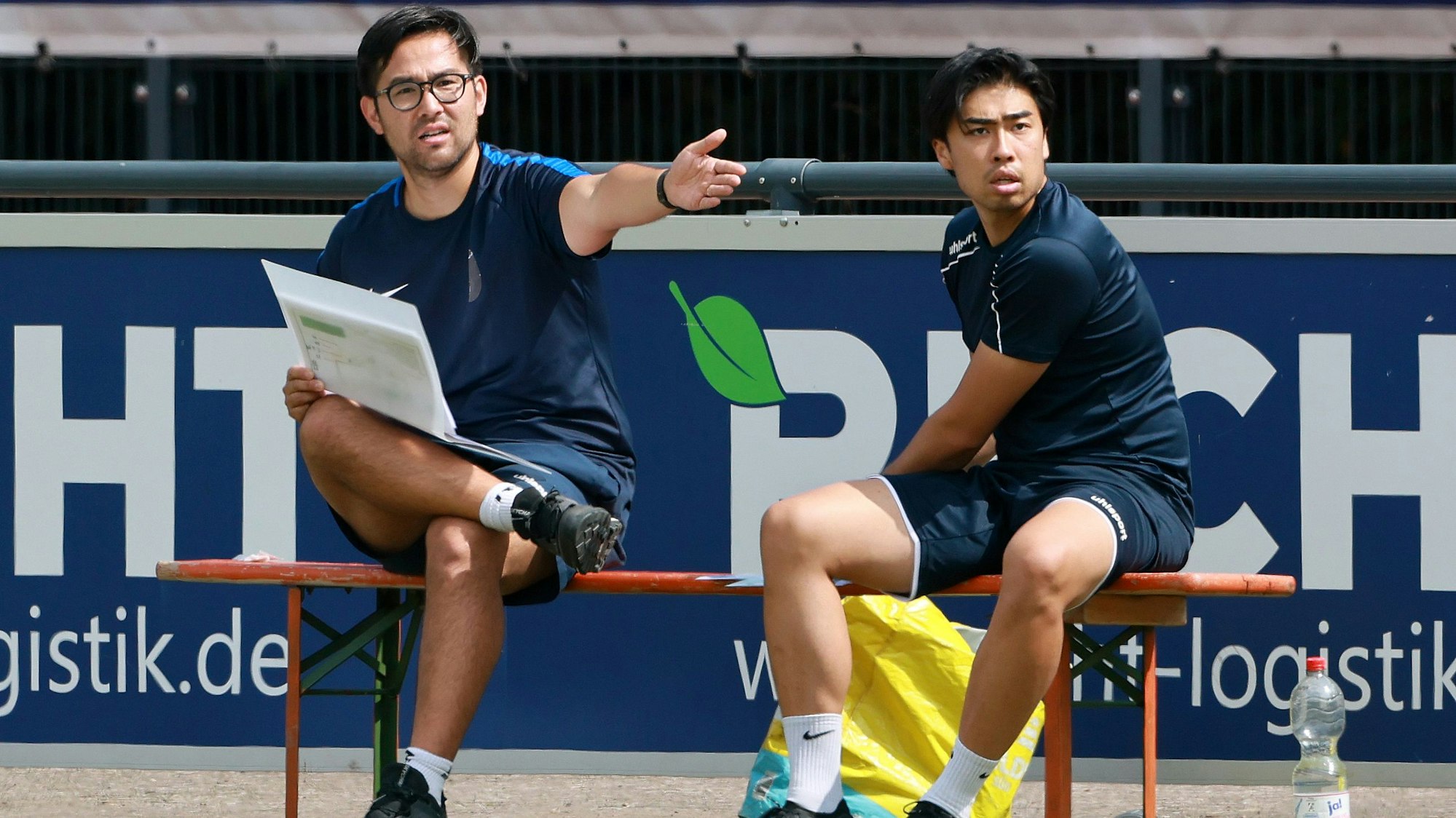 Das Foto zeigt zwei Trainer auf der Bank, der Linke streckt den Arm aus.