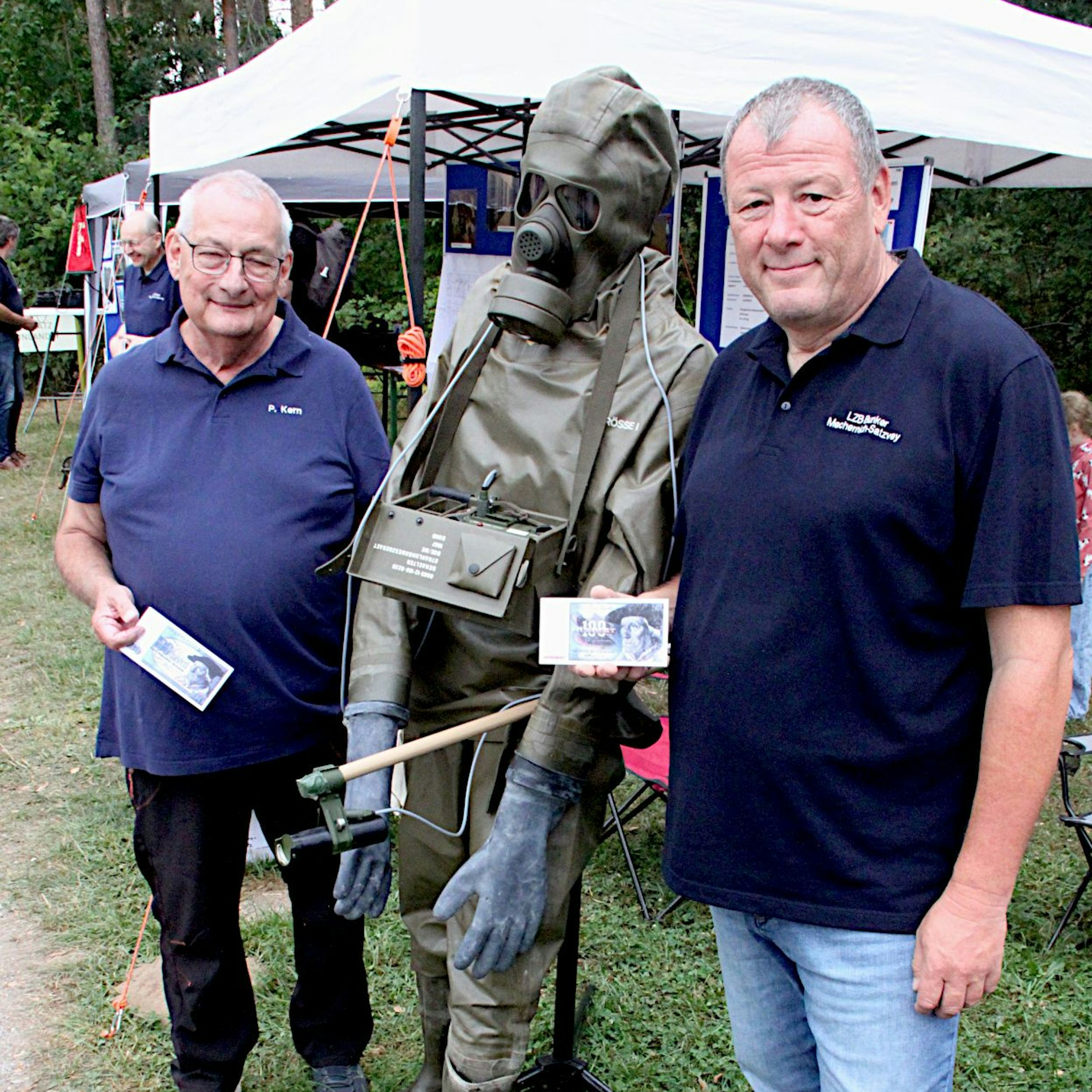 Peter Kern und Lothar Lubitz haben einen Infostand zur Bunkeranlage des „Ausweichsitzes der Landeszentralbank Nordrhein-Westfalen“ aufgebaut. Sie halten einen grünen Vollschutzanzug.