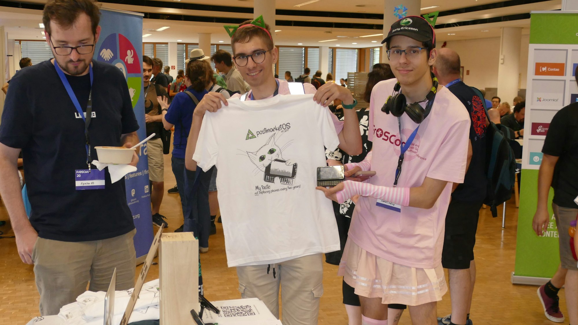 Drei Männer beim Besuch einer Messe. Zwei halten ein T-Shirt mit Aufdruck in die Kamera, einer trägt ein rosa T-Shirt und einen rosa Rock.