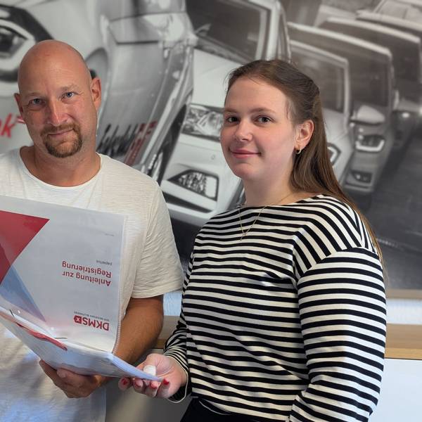 Das Foto zeigt zwei Personen mit einer Infomappe des DKMS vor einem Foto in der Fahrschule Fink in Hürth.