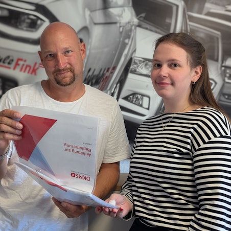 Das Foto zeigt zwei Personen mit einer Infomappe des DKMS vor einem Foto in der Fahrschule Fink in Hürth.