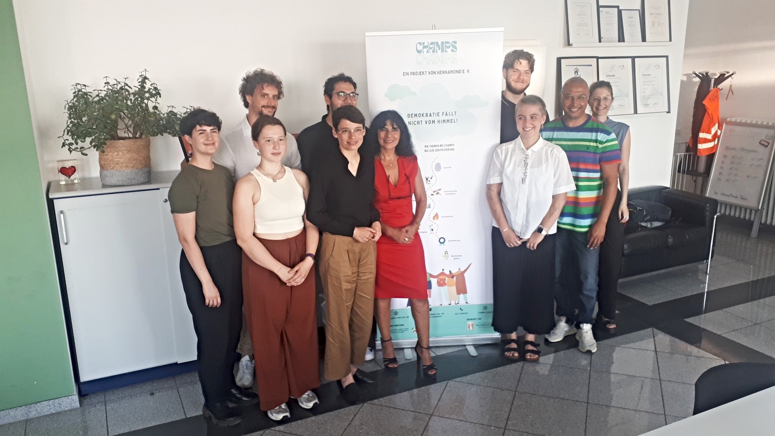 Einige junge Erwachsene und zwei Frauen stehen links und rechts von einem Aufsteller, der das Projekt Champs beschreibt.