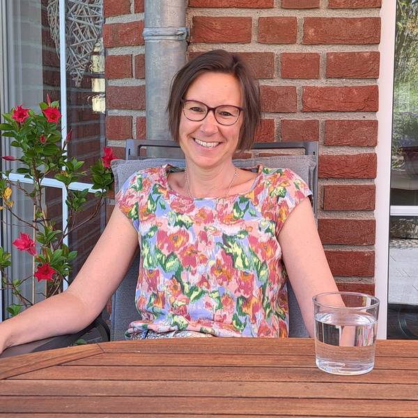 Eine Frau sitzt auf ihrer Terrasse an einem Tisch mit einem Glas Wasser.